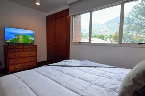 A bedroom with a bed, a TV on a dresser, a window with a view of the outdoors, and a wooden wardrobe. - Tu Rincón Altamira - Nomad Suites (Caracas)