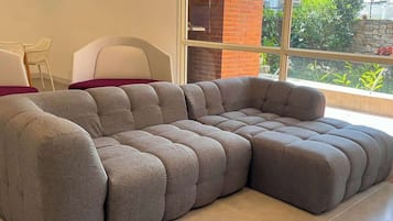 A sectional sofa in a room with a large window, a white chair, and a brick wall.