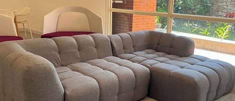 A sectional sofa in a room with a large window, a white chair, and a brick wall.