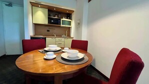 Uma sala de jantar com uma mesa redonda de madeira posta para quatro pessoas, cadeiras estofadas vermelhas e uma área de cozinha com armários e um micro-ondas ao fundo.