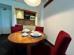 Ein Essbereich mit einem runden Holztisch, gedeckter Platz für vier Personen, roten gepolsterten Stühlen und einer Küchenzeile mit Schränken und einer Mikrowelle im Hintergrund.