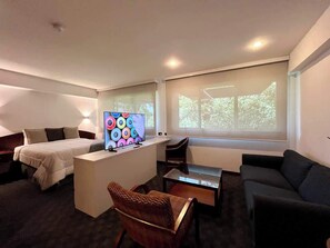 Habitación de hotel moderna con cama, escritorio con computadora, sofá, silla y ventana grande con vistas a árboles.