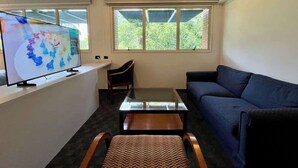 Uma sala de estar com sofá, mesa de centro, televisão em um móvel e janela com vista para vegetação.