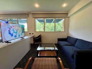 Ein Wohnzimmer mit einer Couch, einem Couchtisch, einem Fernseher auf einem Schrank und einem Fenster mit Blick auf Grünflächen.