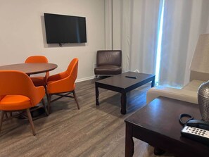Um quarto com televisão na parede, uma poltrona de couro marrom, uma mesinha e um sofá com abajur.