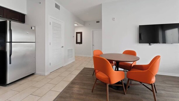 A modern kitchen with a stainless steel refrigerator, a dining area with a round table and orange chairs, and a flat-screen TV mounted on the wall. - 2BR Pool View Suite Near Walt Disney World Resort (Kissimmee)