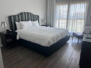 Ein Hotelzimmer mit einem großen Bett, zwei Nachttischen, einem Sessel und einem fenster mit Vorhängen.