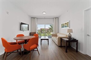 A modern living room with a dining area, a sofa, a TV mounted on the wall, and a balcony with a view. - 2BR Pool View Suite Near Walt Disney World Resort (Kissimmee)