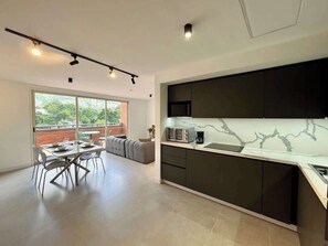 A modern kitchen with dark cabinetry and a marble countertop, a dining area with a table and chairs, and a large window with a view of greenery. - Moderno Ph en Altamira con hermosa vista al Ávila (Caracas)