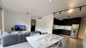 Un soggiorno moderno con angolo ristoro, un divano grigio, una TV a schermo piatto e una cucina con frigorifero.