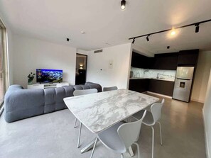 A modern living space with a dining area, a gray sofa, a flat-screen TV, and a kitchen with a refrigerator. - Moderno Ph en Altamira con hermosa vista al Ávila (Caracas)