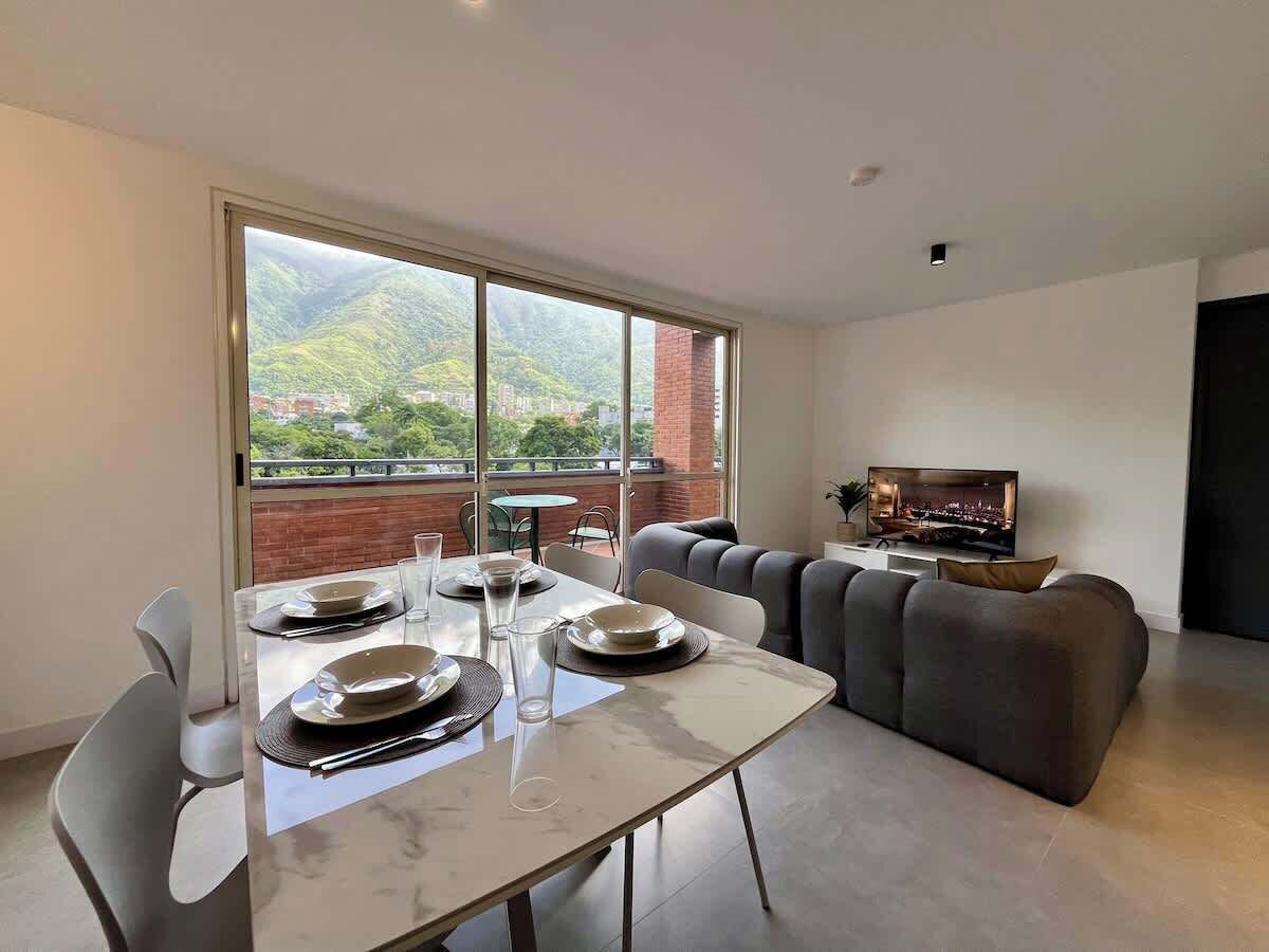 Ein modernes Wohnzimmer mit Essbereich, einer Couch, einem Fernseher und einem Balkon mit Blick auf Grünflächen und Gebäude.