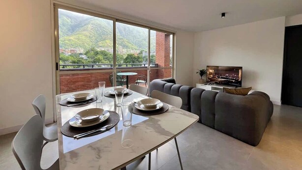 Un soggiorno moderno con zona pranzo, un divano, una televisione e un balcone con vista su spazi verdi ed edifici.