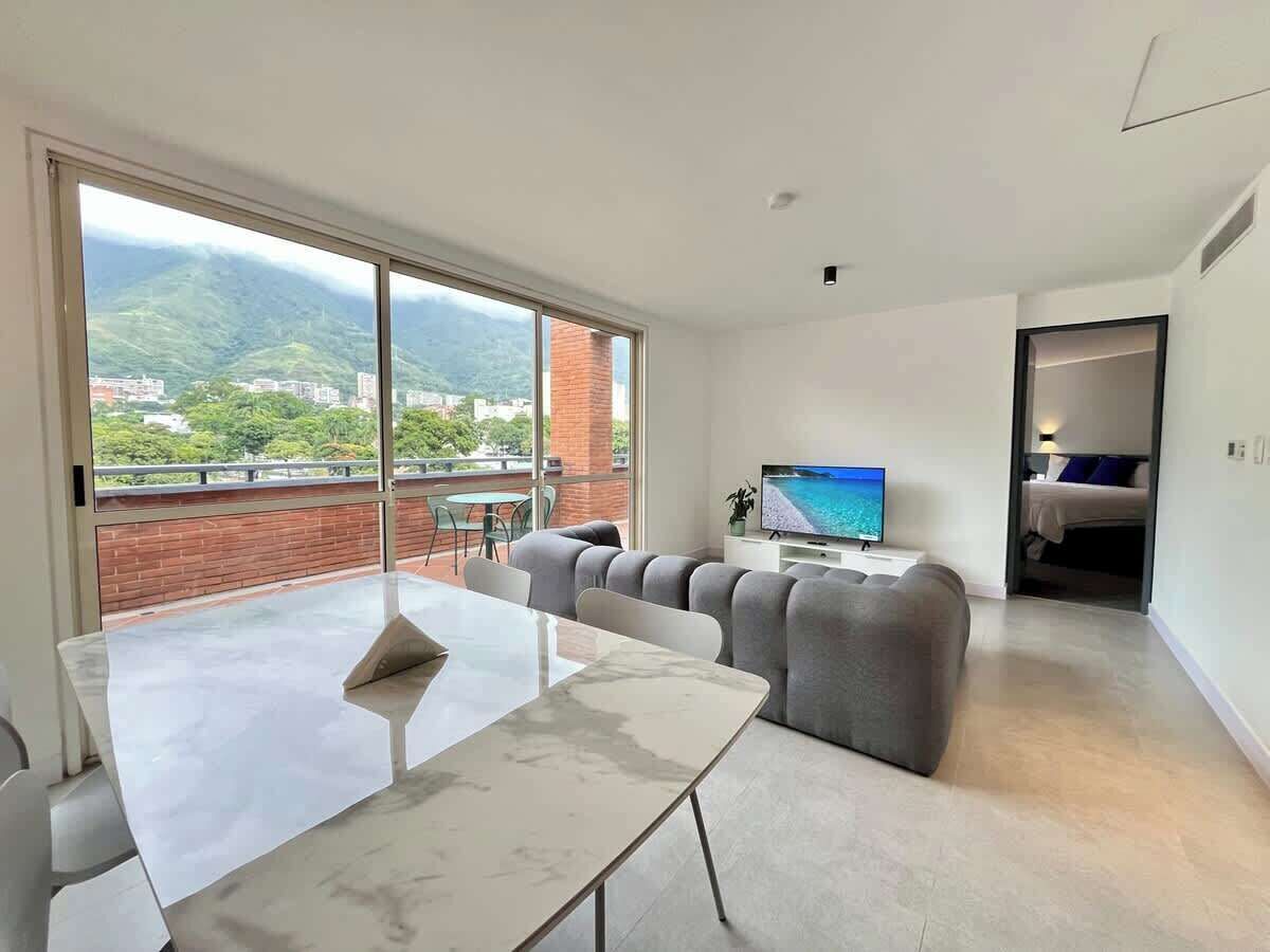 A modern living room with a large window, a flat-screen TV, and a dining area.