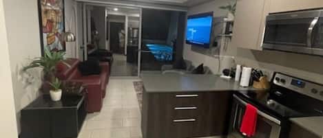 A modern kitchen with a central island, built-in microwave, and oven. There is a dining area with a sofa and a TV.
