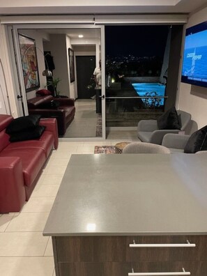 Una sala de estar moderna con un televisor grande, comedor y vista a una piscina por la noche.