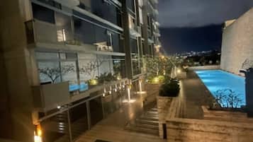 A nighttime view of a modern building with multiple balconies, a swimming pool, and outdoor lighting.