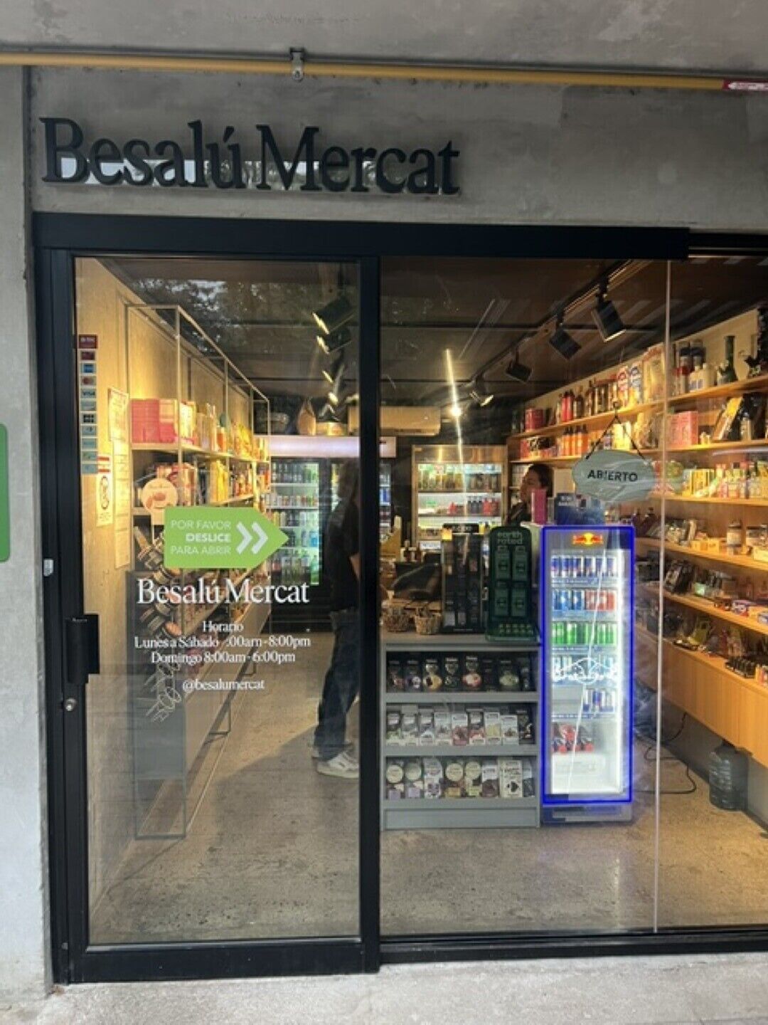 Un magasin appelé « Besalú Mercat », avec des étagères remplies de divers produits, un rayon réfrigéré et un panneau indiquant que le magasin est ouvert.