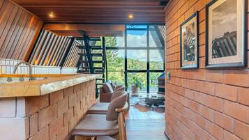 A bar area with a wooden counter and seating, a large window with a view of greenery, and a brick wall with framed pictures.