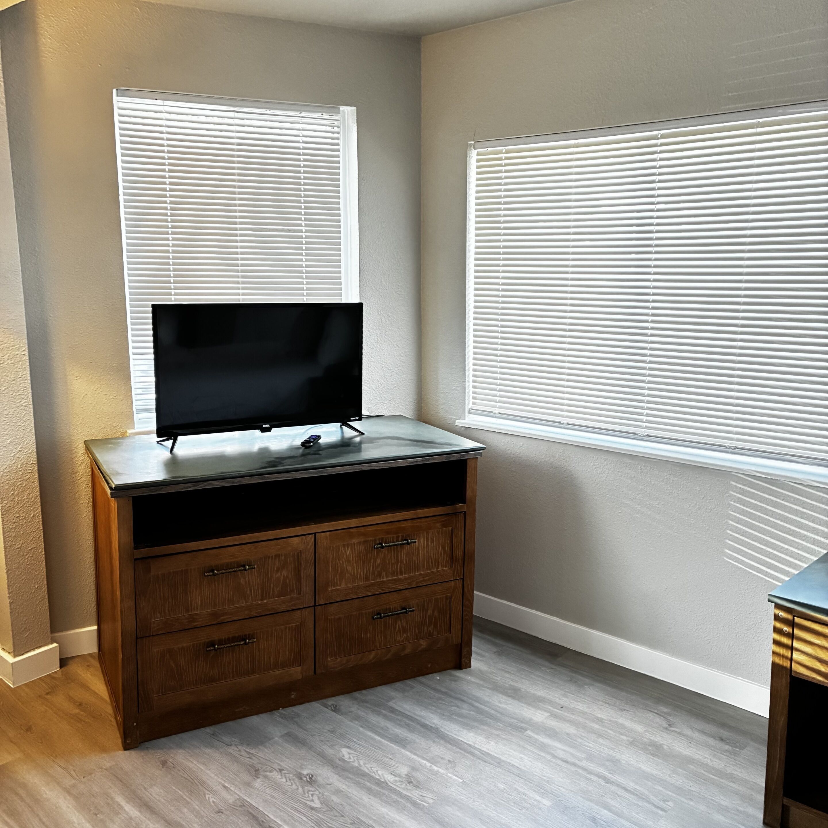 A room with a TV on a wooden dresser, two windows with blinds, and a light-colored floor.