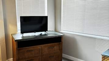 A room with a TV on a wooden dresser, two windows with blinds, and a light-colored floor.