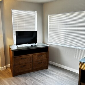Una habitación con televisor sobre un aparador de madera, dos ventanas con persianas y un suelo de color claro.
