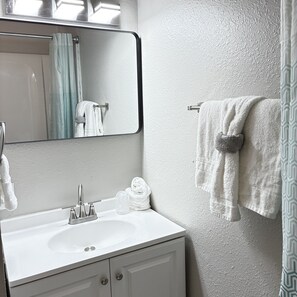 Un baño con lavamanos blanco, espejo, toallero con toalla y cortina de ducha.