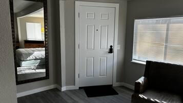 A room with a white door, a large mirror, a window with blinds, a dark-colored chair, and a bed with white bedding.