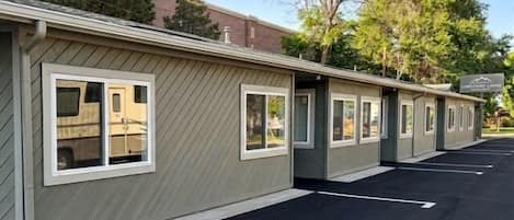 A row of motel-style buildings with individual rooms, a parking lot, and trees in the background.
