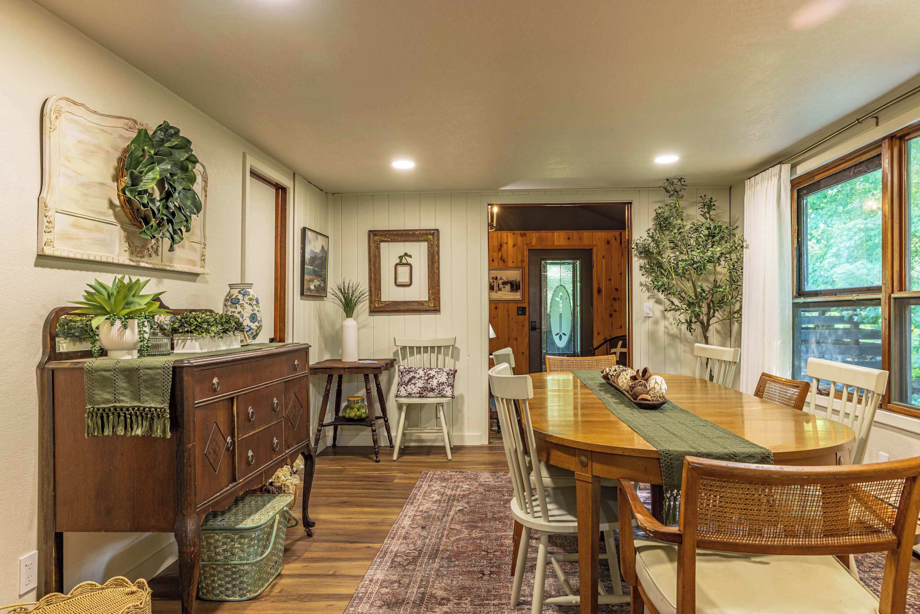 A dining area with a wooden table, chairs, and a rug. There are plants and decorative items on the walls and a wooden door leading to another room.