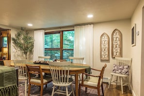 A dining area with a round table, six chairs, and a view of greenery through large windows.