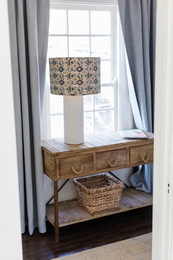 Un escritorio de madera con una lámpara, una cesta y una ventana con cortinas.