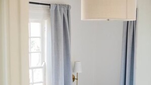 Un dormitori amb un llit, una tauleta de nit, una làmpada i una finestra amb cortines.