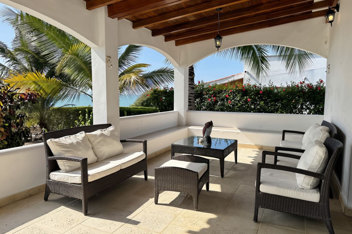 A covered outdoor seating area with wicker furniture and a glass table, surrounded by white walls and tropical plants.