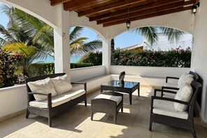 Un espacio de estar al aire libre cubierto con mobiliario de mimbre y una mesa de vidrio, rodeado de paredes blancas y plantas tropicales.