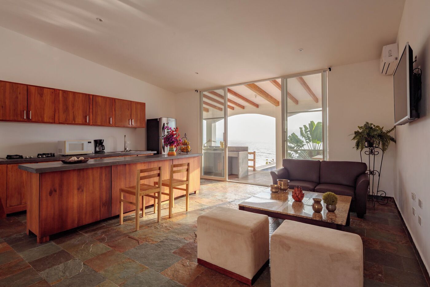 A modern kitchen with wooden cabinets, a central island with bar stools, and a dining area with a sofa and coffee table.