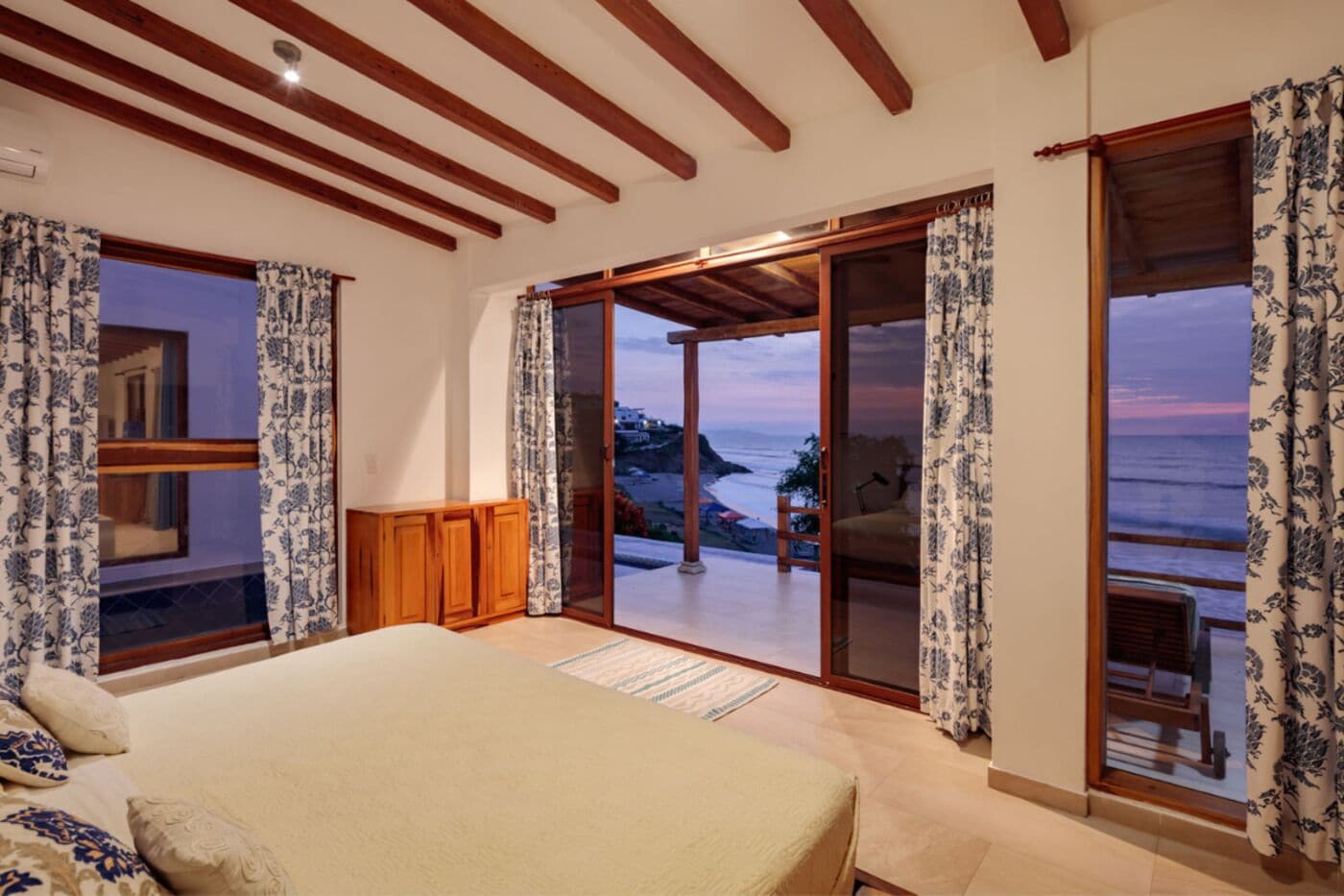A bedroom with a bed, a wooden cabinet, and a view of the sea through a sliding glass door.