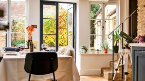 A dining area with a table set for two, a telescope on a tripod, and a view of a garden through large windows. - Cotswolds Manor With Garden (Lechlade)