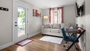 A compact living space with a sofa, a desk with a laptop, a small rug, and a door leading outside. - Lemon Door Cottage (St. Petersburg)