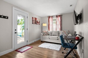 Ein gemütlicher Wohnbereich mit Sofa, Schreibtisch mit Laptop, kleinem Teppich und einer Tür, die nach draußen führt.