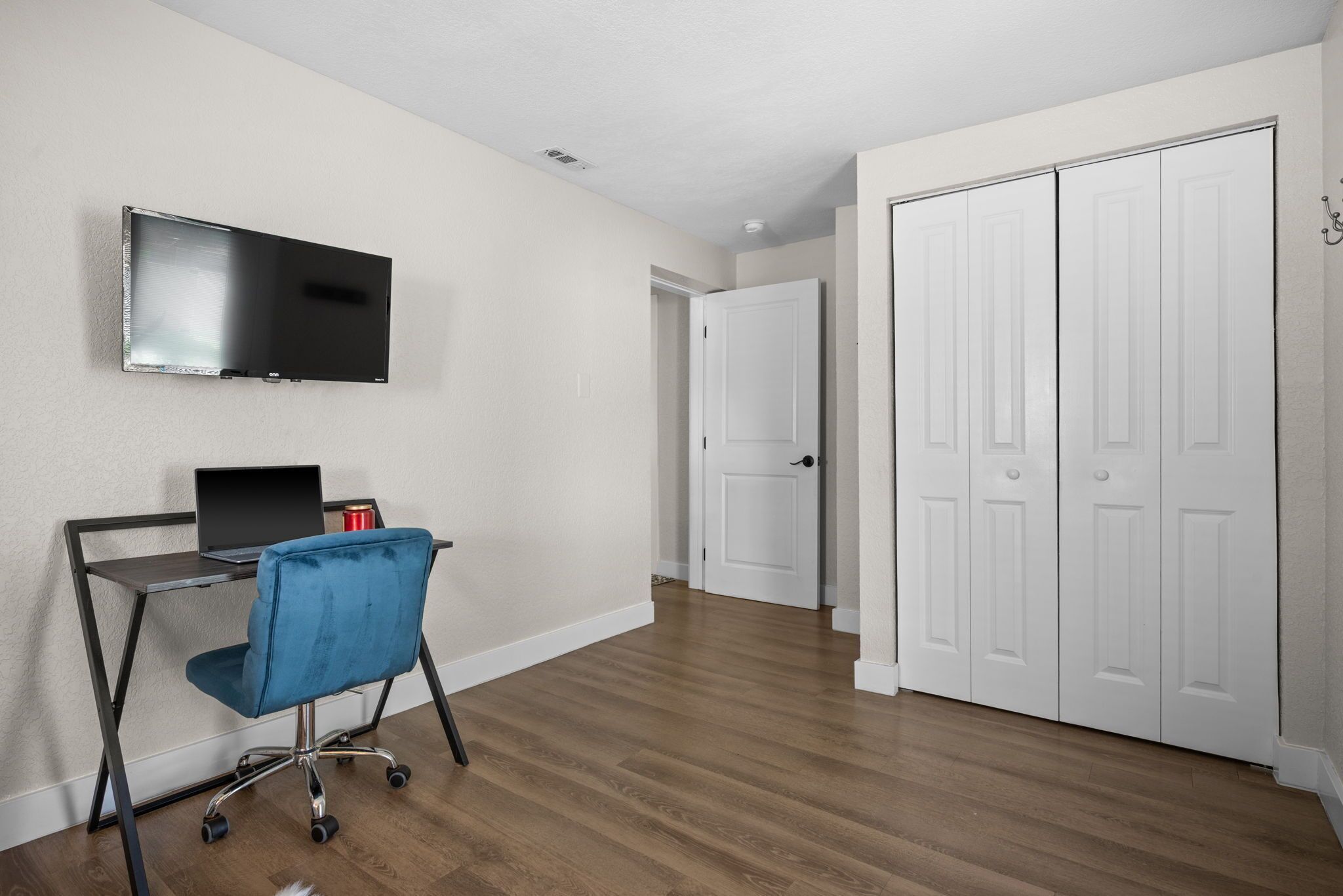 A room with a flat-screen TV mounted on the wall, a desk with a laptop and a blue chair, and a closed white door.