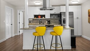 A modern kitchen with white cabinetry, a stainless steel range hood, a built-in oven, a refrigerator, and two yellow bar stools. - Lemon Door Cottage (St. Petersburg)