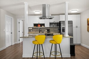 Eine moderne Küche mit weißen Schränken, einer Edelstahl-Dunstabzugshaube, einem Backofen, einem Kühlschrank und zwei gelben Barhocker.