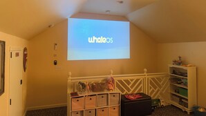 Ein Zimmer mit Deckenprojektor, der 'whalooos' zeigt, einer weißen Stauraumbank mit Schubladen und einem kleinen Spielzeug-LKW auf dem Boden.