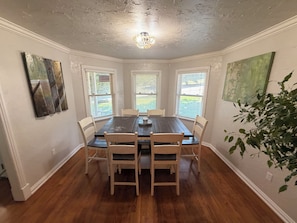 A dining area with a wooden table and chairs, three windows with blinds, and a chandelier.