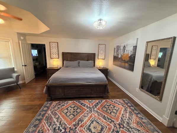 A bedroom with a large bed, two bedside tables with lamps, a patterned rug, a cityscape painting, and a mirror.
