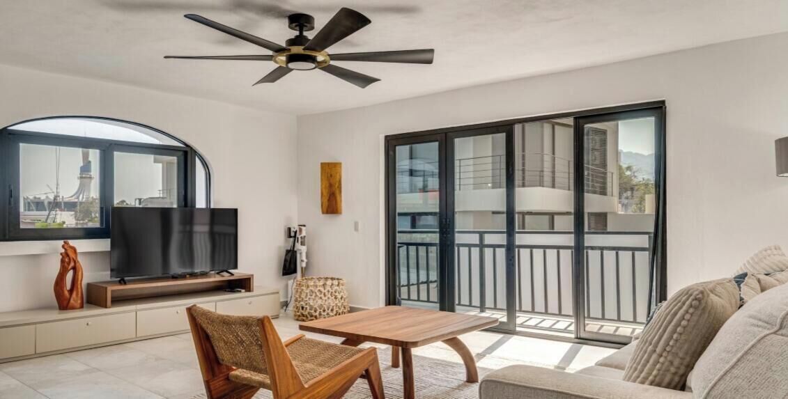 Ein modernes Wohnzimmer mit Flachbildfernseher, Deckenventilator, einer Couch, einem Couchtisch und einer großen Glasschiebetür, die auf einen Balkon führt.