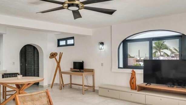 A modern living room with a ceiling fan, a TV on a wooden stand, a dining table with chairs, and a large window with a view. - Modern Oasis 2BR Condo/ Walk to Camarones Beach & Malecón (Puerto Vallarta)