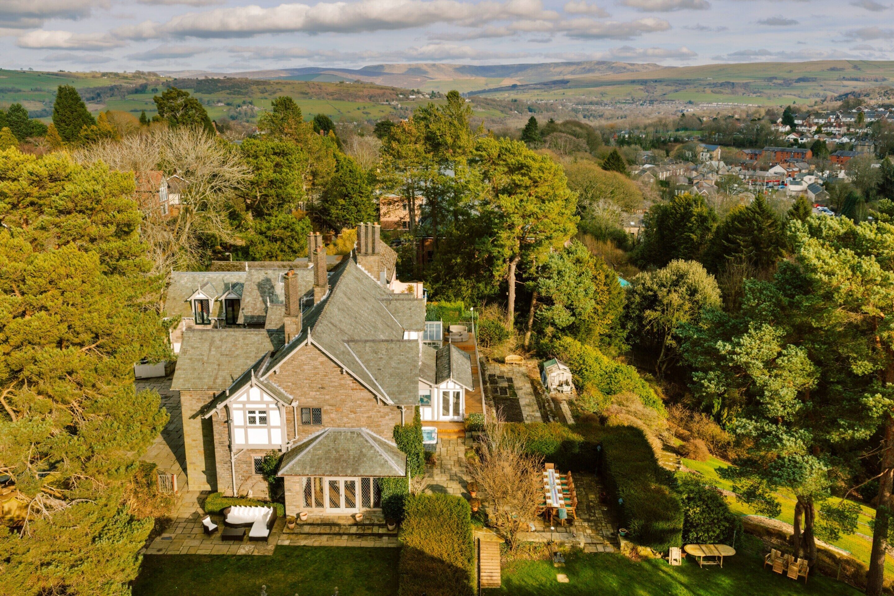 Une grande maison dotée d’un toit à pignon imposant, entourée d’une végétation luxuriante et offrant une vue sur une ville au loin.