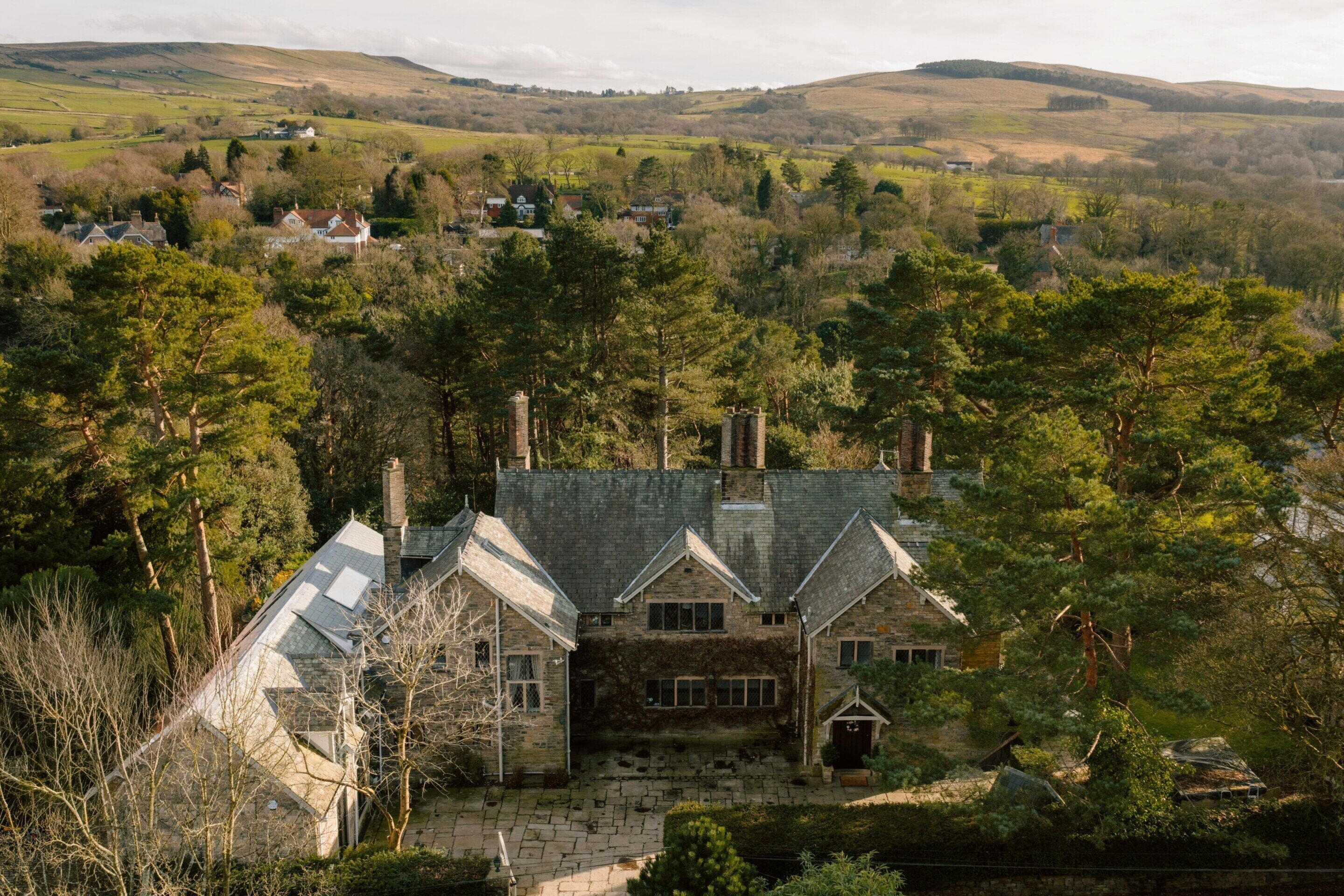 A stone house with a steep roof, surrounded by dense trees and a hilly landscape in the background.